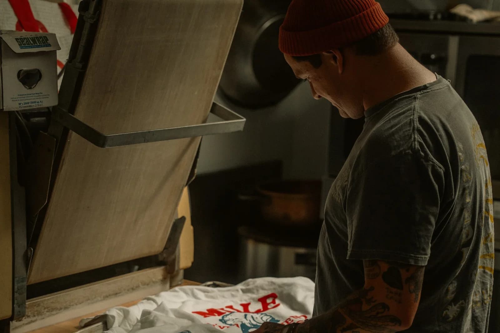 An operator inspecting a printed white T-shirt beside an open industrial heat press